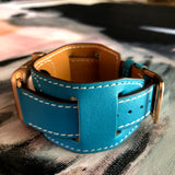 Cyan Blue Apple Watch Leather Cuff by Juxli Home.  Handmade, stylish leather strap with rose gold hardware on a 40mm Apple watch on a canvas with a black and gray painting.